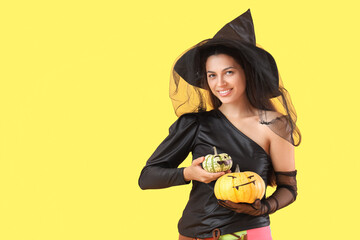 Young woman dressed for Halloween as witch with pumpkins on yellow background