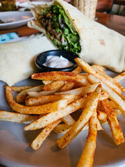 A plate of french fries and a shawarma with a black container of sauce. The shawarma and the fries are seasoned