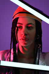 Beautiful young African-American woman with braids holding neon lamps on purple background
