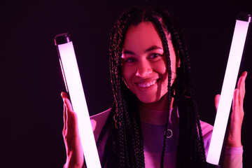 Happy young African-American woman with braids holding neon lamps on dark background