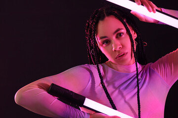 Beautiful young African-American woman with braids holding neon lamps on dark background