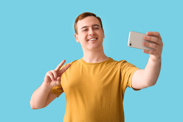 Young man with mobile phone showing victory gesture and taking selfie on blue background