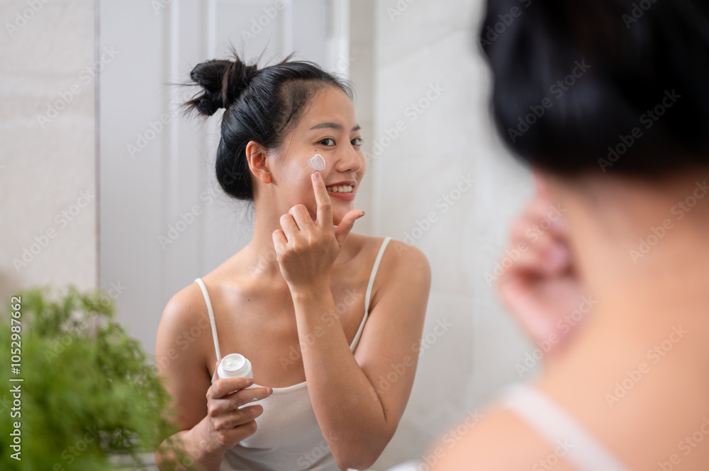 Wall mural A natural Asian woman is applying facial cream in front of the mirror in the bathroom after shower.