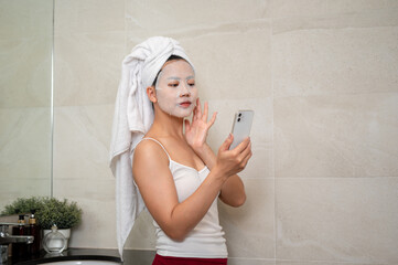 An Asian woman with a facial treatment mask on her face is using her smartphone in the bathroom.