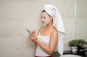 An Asian woman with a facial treatment mask on her face is using her smartphone in the bathroom. - Powered by Adobe