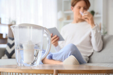 Filter jug on table against young man with tablet computer drinking water at home, closeup