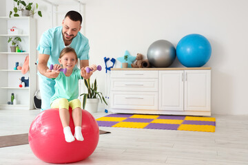 Male physiotherapist and little girl with dumbbells training on fitball in rehabilitation center