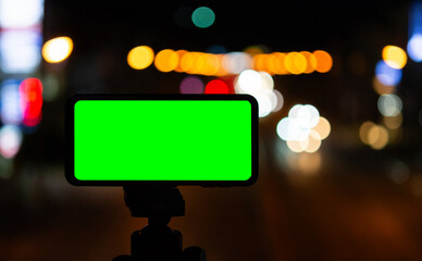 Smart mobile phone with green screen placed on a tripod with blurry background bokeh of night lights.