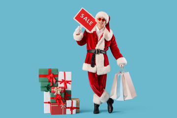 Young Santa Claus with SALE sign and shopping bags near Christmas presents on blue background