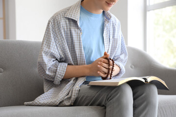 Teenage boy with prayer beads and Holy Bible sitting on sofa at home, closeup