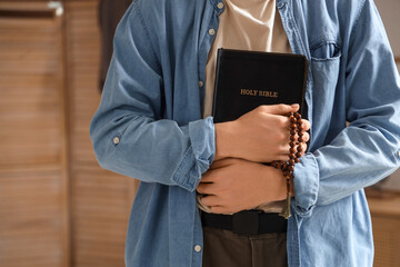 Teenage boy with Holy Bible and prayer beads in bedroom, closeup