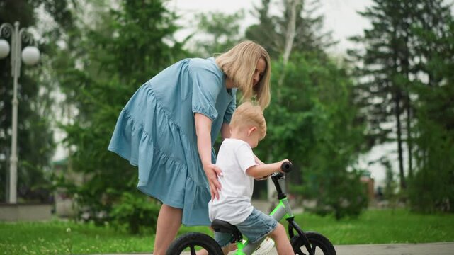 A caring mother is gently guiding her focused young son as he rides his bicycle along an interlocked pathway, she touches his head to offer support and help him maintain balance