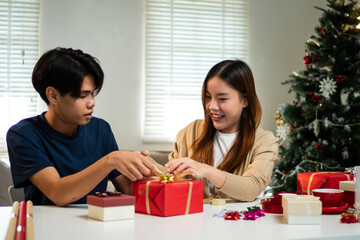 A young man and woman are sitting together at a table, wrapping gifts in a cozy, festive setting. A...