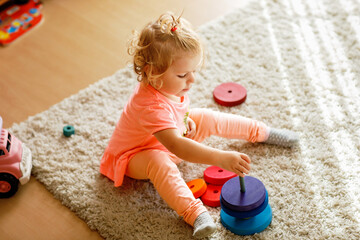 Happy toddler girl sits on a carpet, playing with colorful wooden rings and a toy truck, focused on stacking the pieces. Child plays with rainbow colored pyramid at home or nursery.
