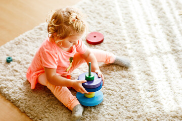 Happy toddler girl sits on a carpet, playing with colorful wooden rings and a toy truck, focused on stacking the pieces. Child plays with rainbow colored pyramid at home or nursery.