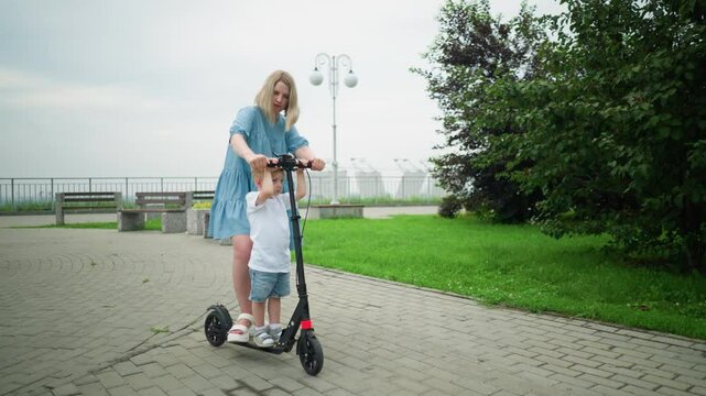 A mother and her young son stand together on an electric scooter, on an interlocked pathway