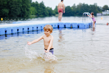 Little blond preschool boy having fun with splashing in a lake on summer day, outdoors. Happy child learning swimming. Active leisure with kids on vacations. Danger on domestic lakes