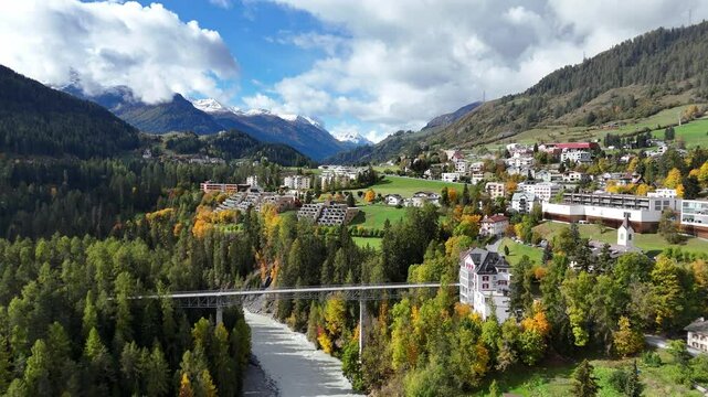 A scenic swiss village surrounded by mountains, forests, and a river below, aerial view