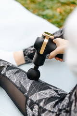 Fototapeta premium young attractive fitness woman in sportswear with athletic build makes self massage with percussion gun, sitting on mat on green grass in park, legs massage, cropped, unrecognizable