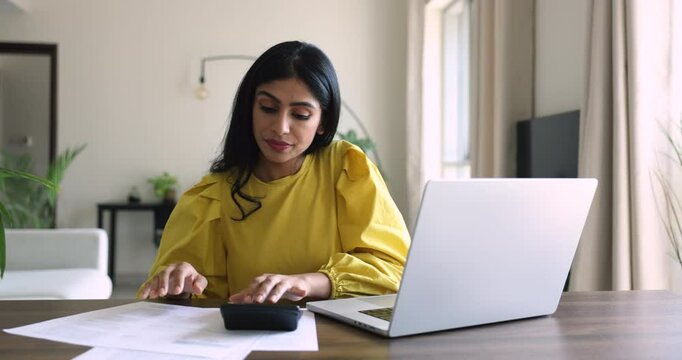 Busy young Indian freelance accountant woman working at home, typing on laptop, using calculator, counting budget, expenses, fees, taxes, consulting paper documents, reports, invoice