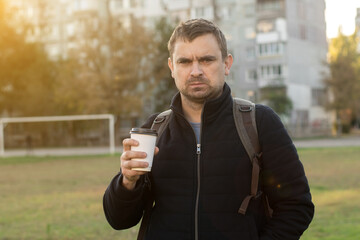 Man at a stadium with coffee cup and backpack, captured in an emotional moment. Background of high-rise buildings under evening light. 