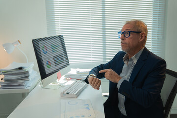 Senior manager pointing at his wristwatch while looking away, sitting at desk with computer and...
