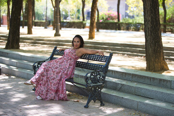 a young, beautiful brunette woman is sitting on a park bench, her arms resting on the backrest, her...