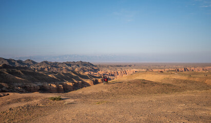 An expansive desert vista unfolds under a vast sky, where travelers traverse the arid landscape of rolling hills and layered canyons, with distant snow-capped mountains providing a majestic backdrop.