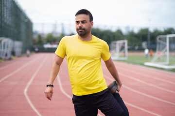 Energetic athlete in wireless headphones prepares leg muscles for training on rubberized running track at urban park's sports ground.