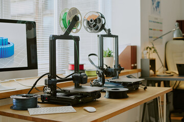 Dual 3D printers operating on wooden desk beside computer displaying object model. Office environment featuring various tech gadgets and decor pieces