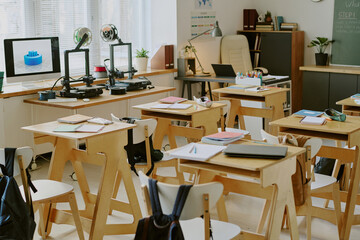 Classroom setting with multiple 3D printers on tables, organized desks with notebooks and stationery, sunlight streaming through windows, and blackboard in background