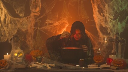 A witch in black clothing conjures up a skull sitting in a scary kitchen with pumpkins and potions.