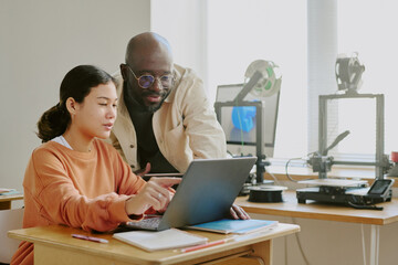 Hispanic woman working with African American man on laptop in well-lit modern office equipped with 3D printer and model replicating space, showing professional engagement