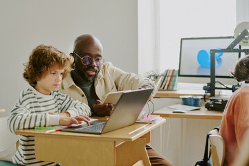 African American teacher mentoring focused young student using laptop in bright classroom. Books...