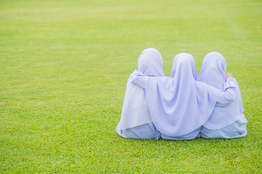 Three girls wearing white hijabs sit hugging each other on a green grass background.