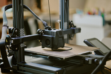Close-up of a 3D printer during operation in a workshop setting creating an object with detailed components visible and equipment in the background.
