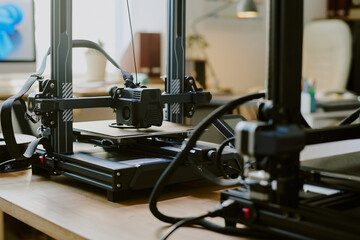 Three-dimensional printing device on wooden desk in well-lit office with various pieces of equipment and decor visible in background