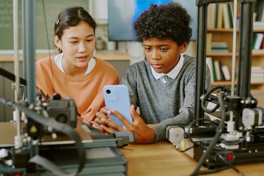 Two students observing a 3D printer's progress in a classroom setting involving technology education. Close-up view of their focused expressions showing concentration around printer setup equipment