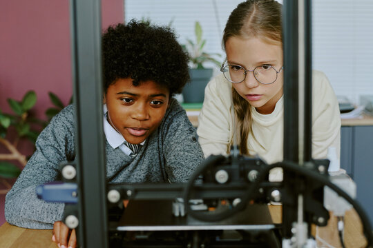 Young boy and girl focused on learning 3D printing technology together. They are concentrating on the 3D printer in classroom environment with plants and desk background