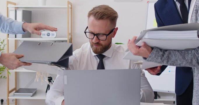 Stressed man sitting at the desk in office frustrated and overwhelmed by coworkers at workplace. Young businessman tired from colleagues. Overworked, multitasking concept. 4k video. Slow motion video