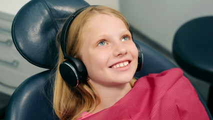 Smiling girl wearing headphones at a dental clinic. A young girl is sitting in a dental chair,...