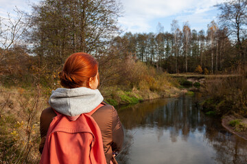 Back view of woman with backpack walking in wildlife national park. Traveler standing on river bank. Concept of ecotourism, hiking, travel in autumn.