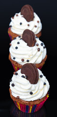 Line up of three frosted cupcakes on black plate with white cream and brown biscuits