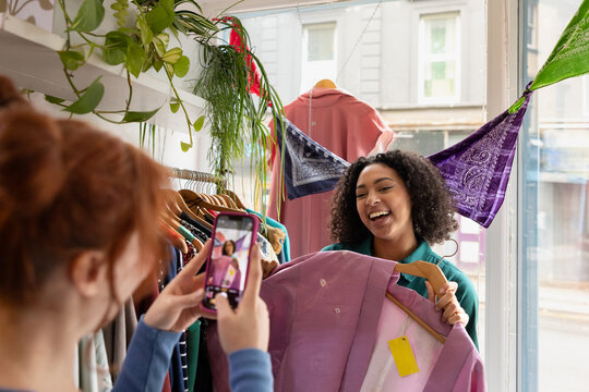 Female friends shopping in a thrift store having fun trying on clothes and taking photos with a cell