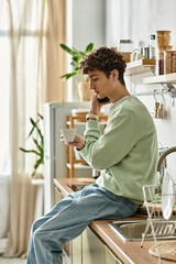 Curly-haired young man relaxes in a contemporary kitchen, sipping coffee and engaged in...