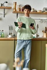 In a contemporary kitchen, a young man with curly hair reviews his notes and uses his phone for...