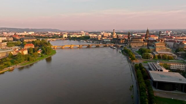 Drone video of Dresden during a flood with warm light illuminating the scene. A tram crosses a bridge, while the damaged Carola Bridge is visible in the background. Drone move forward.