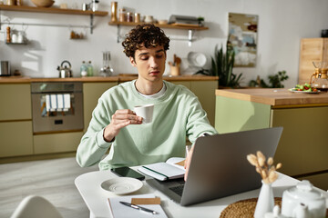 A handsome young man sips coffee from a cup while focused on his laptop in a contemporary home...