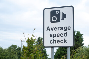 Large roadside sign notifying motorists, that the stretch of highway ahead is an Average Speed Check zone. Low view of sign against a clear sky with green bushes nearby.