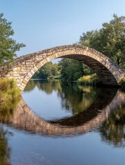 Fototapeta premium A stone arch bridge reflected in calm waters, surrounded by lush greenery and a clear sky.